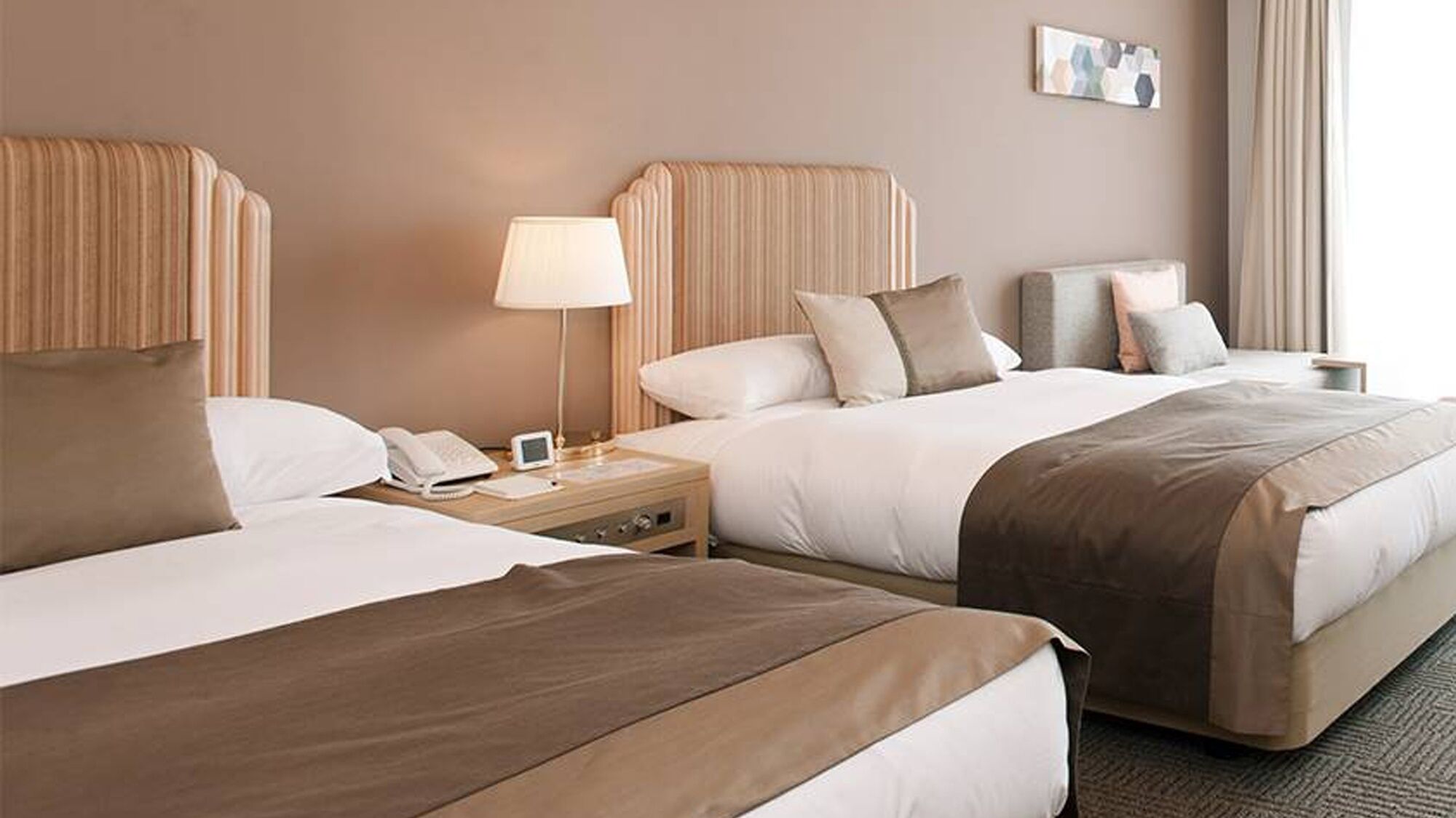 A hotel room with two beds, a bedside table with a lamp, a phone, and a small screen.