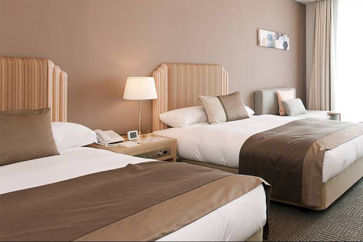 Quarto de hotel com duas camas, uma mesinha de cabeceira com abajur, telefone e uma pequena mesa com um livro.