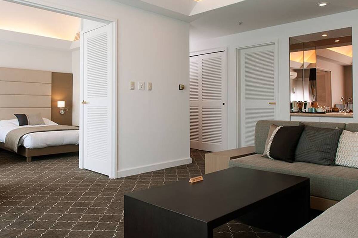 Quarto de hotel com cama, sofá, mesa de centro e espelho refletindo uma cozinha compacta.