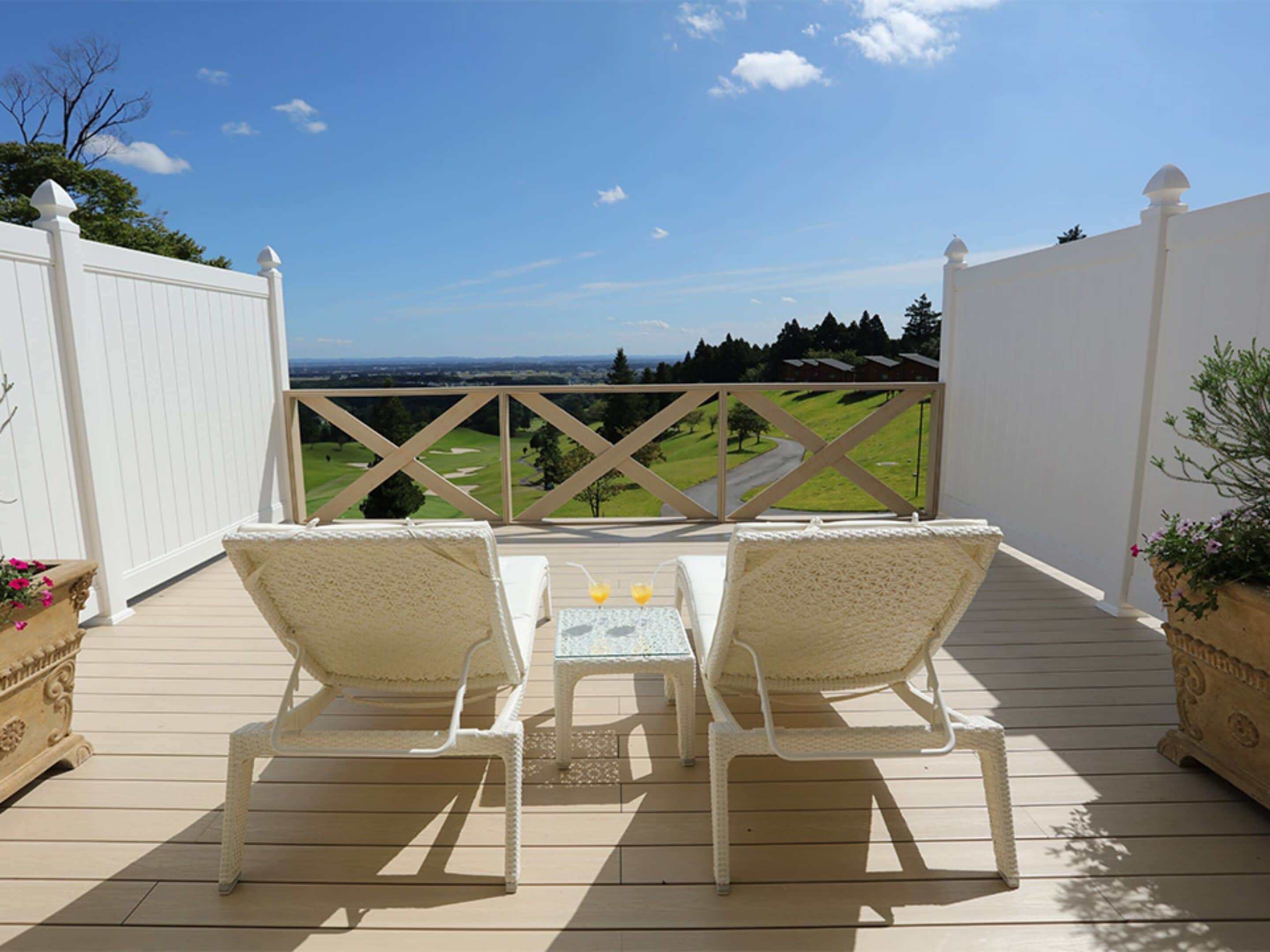 Terraza con dos sillones blancos de mimbre y una pequeña mesa, con vistas a un campo de golf y un paisaje lejano.