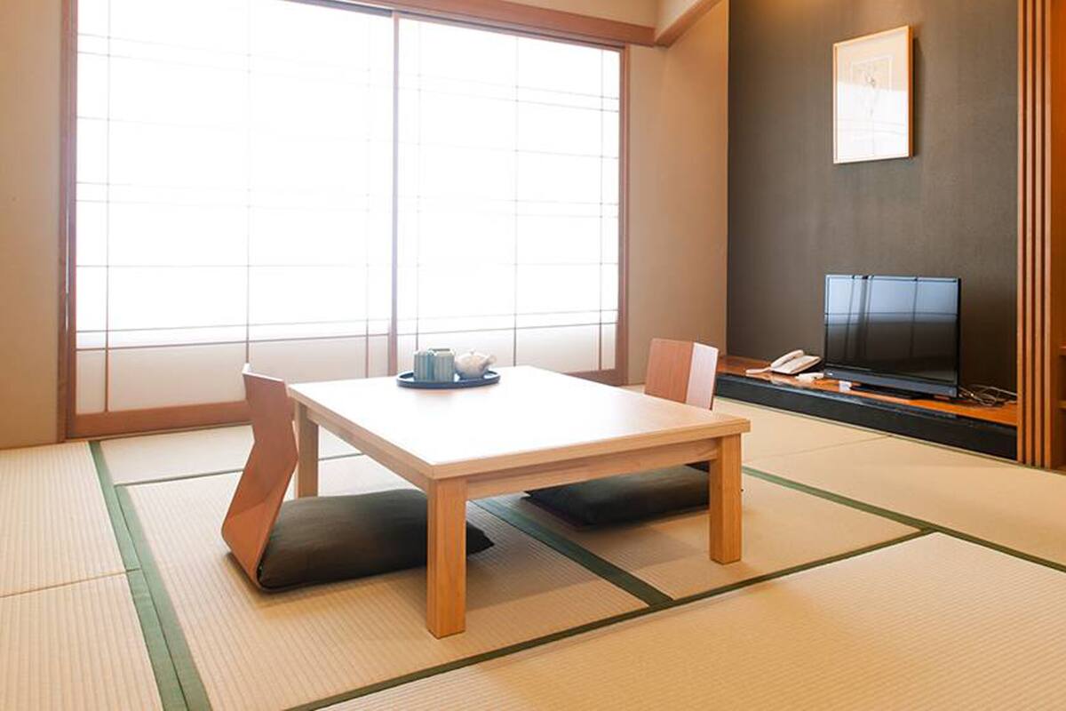 Um quarto japonês tradicional com tatames, uma mesa baixa com almofadas, uma televisão e uma grande janela.