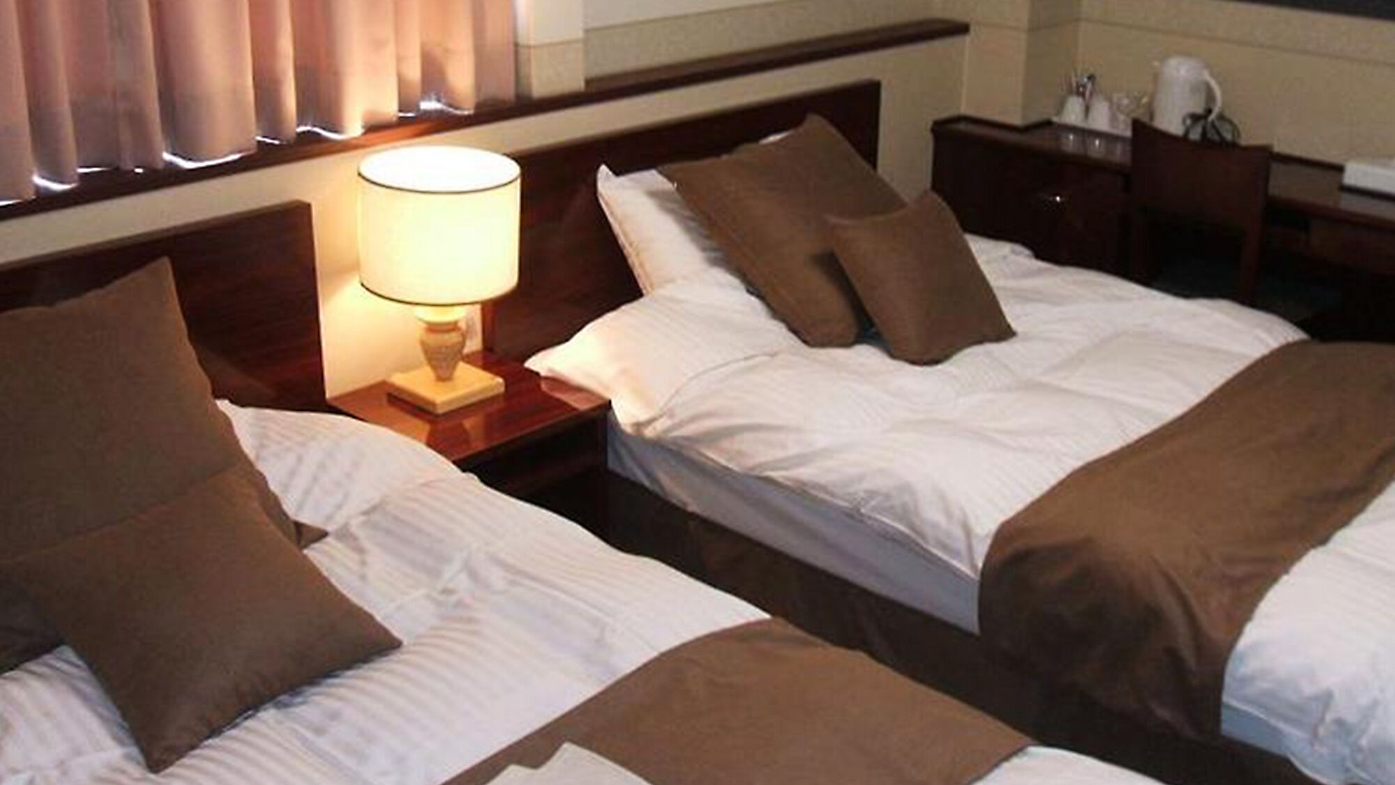 A hotel room with two beds, a nightstand with a lamp, and a dresser with toiletries.
