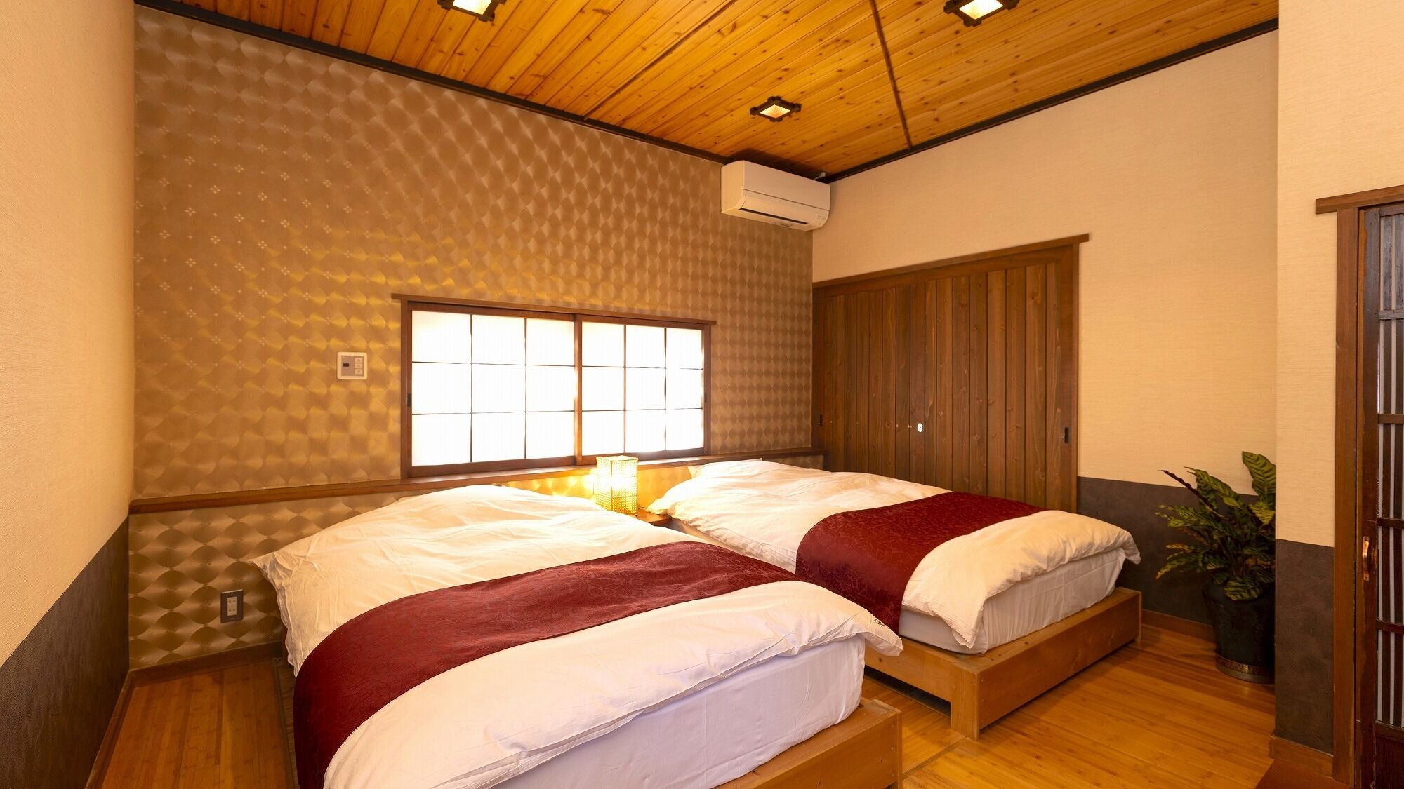 A room with two beds, a wooden ceiling, a window with a view, and a plant.