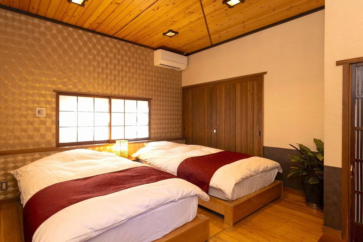 A room with two beds, a wooden ceiling, a window with a view, and a plant.