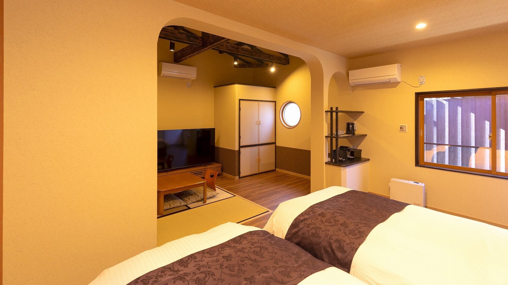 A hotel room with a TV, a bed, a desk, a chair, a small table, a shelf with a coffee maker, and a window.