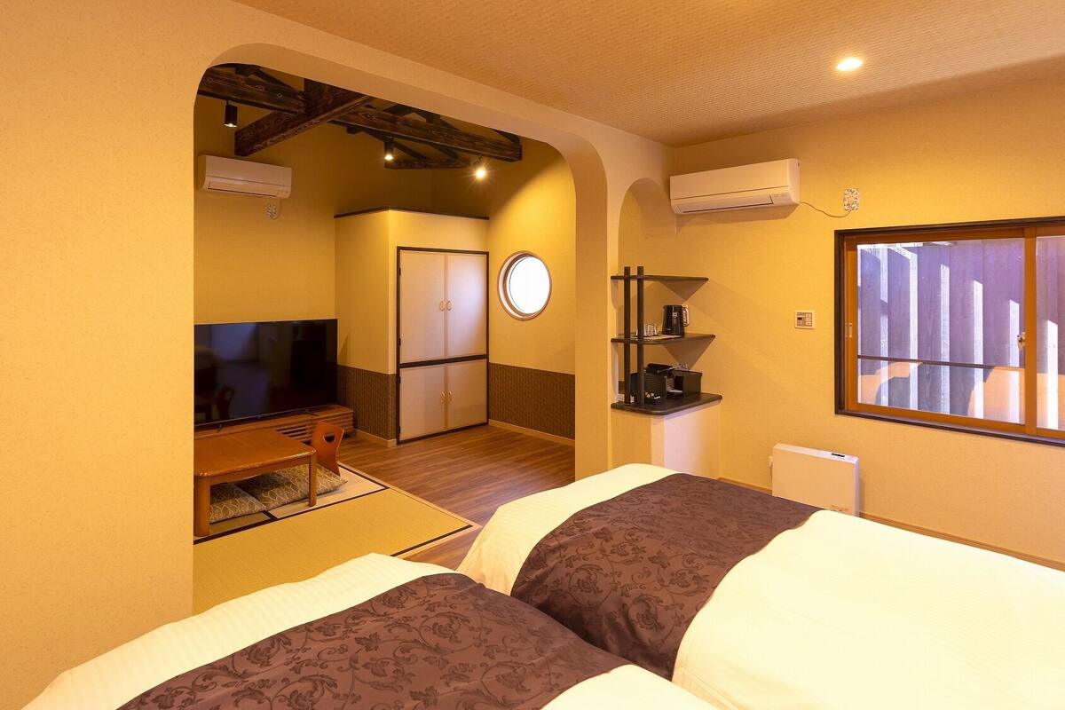 A hotel room with a TV, a bed, a desk, a chair, a small table, a shelf with a coffee maker, and a window.