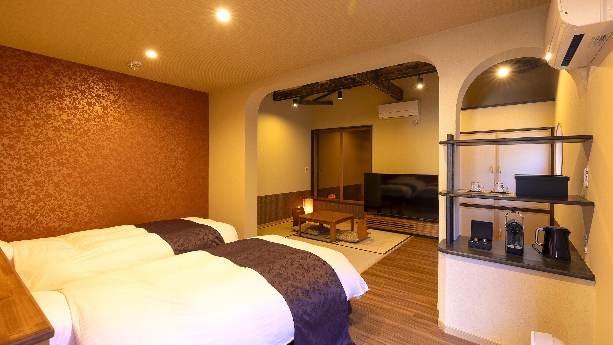 A hotel room with two beds, a small table, a TV, and a kitchenette.