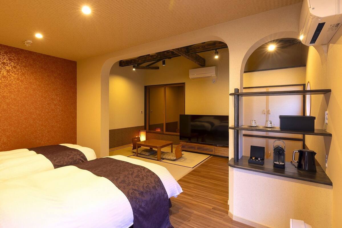 A hotel room with two beds, a small table, a TV, and a kitchenette.
