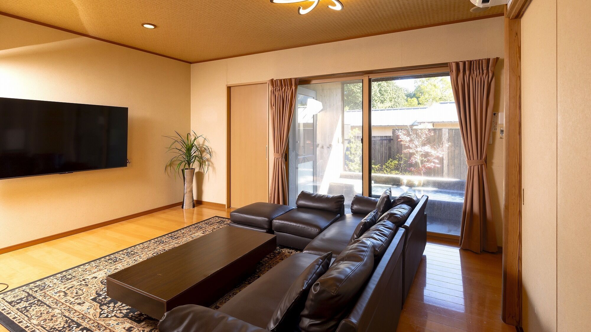 A living room with a sofa set, a flat-screen TV, a potted plant, and a sliding glass door leading to an outdoor area.