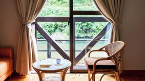 A room with a glass door leading to a view of a bridge and greenery, a wicker chair, a small table with a cup and book, and a sofa. - Hosenbo Lodge (Seiyo)
