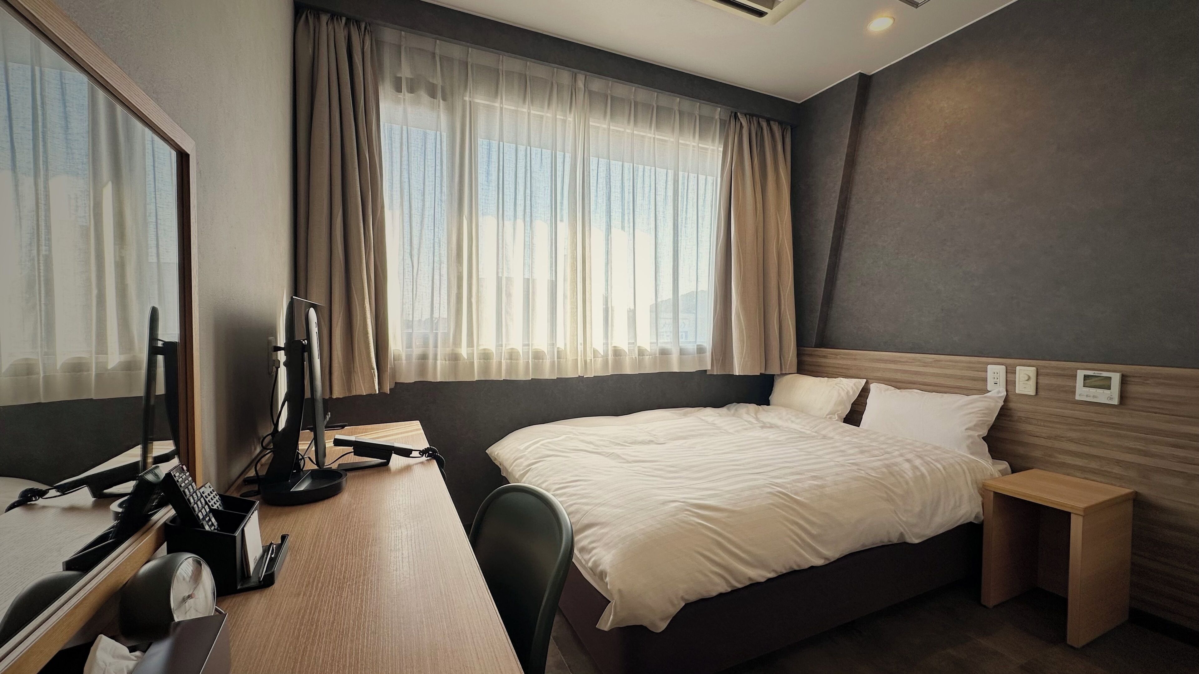 A hotel room with a large bed, a desk with a computer, a chair, and a window with curtains.