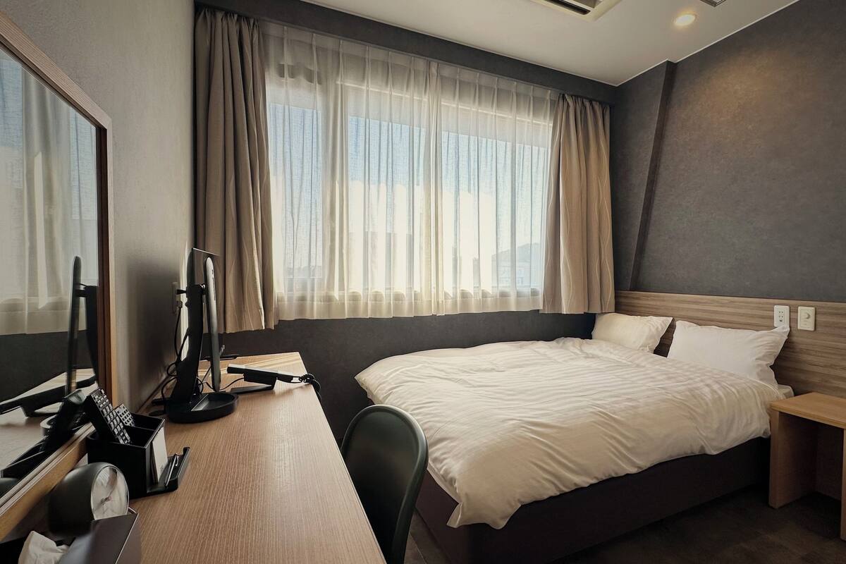 A hotel room with a large bed, a desk with a computer, a chair, and a window with curtains.