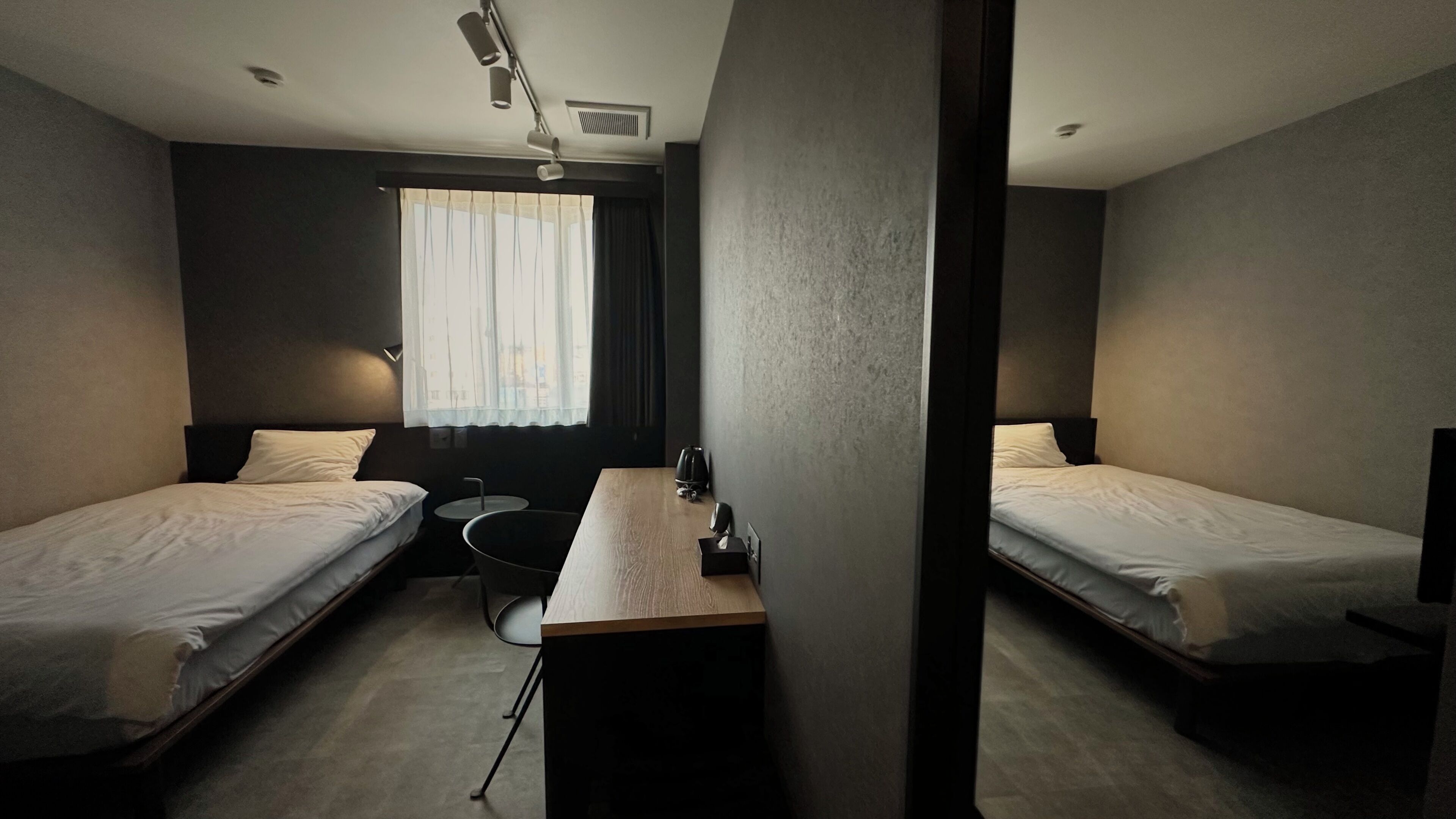A hotel room with two single beds, a small desk, a chair, a window with curtains, and a ceiling vent.