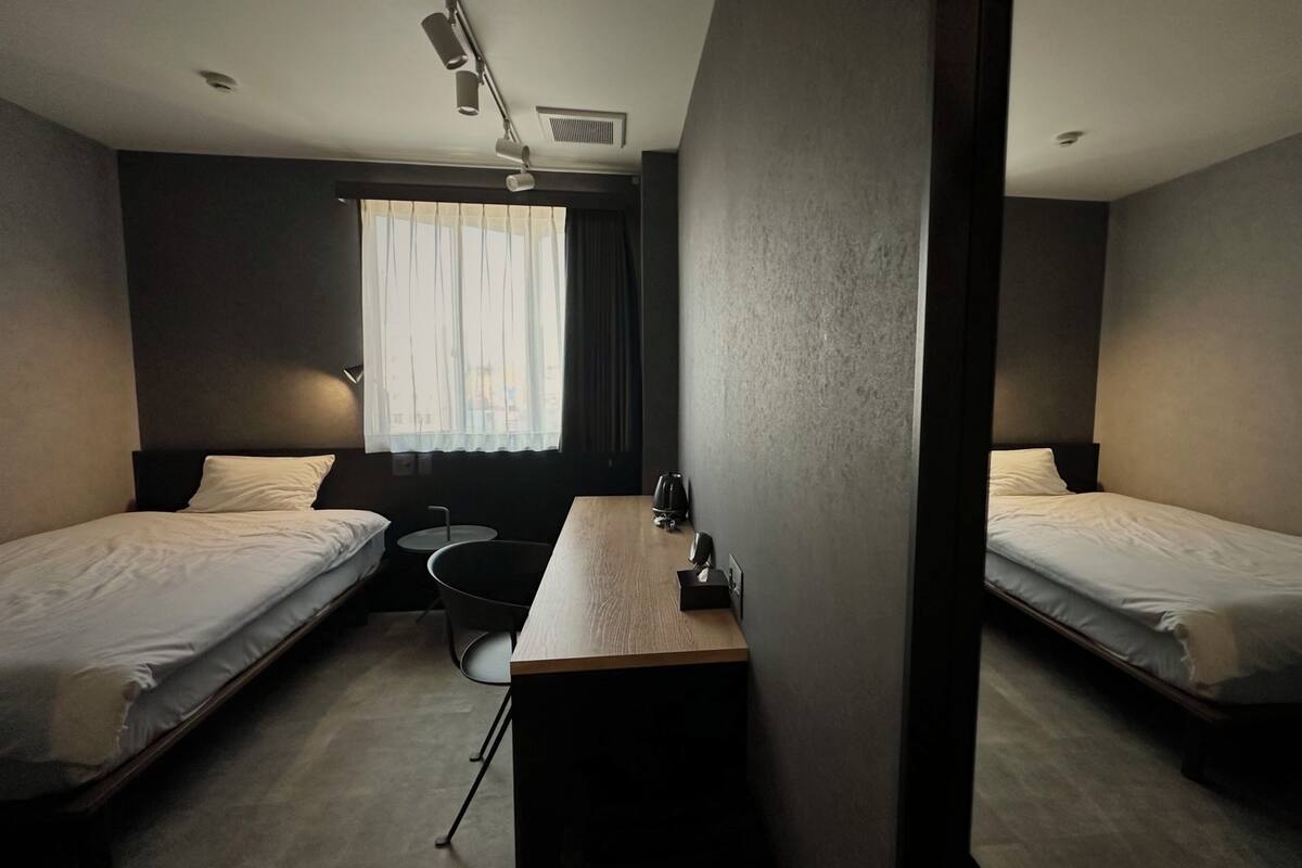 A hotel room with two single beds, a small desk, a chair, a window with curtains, and a ceiling vent.