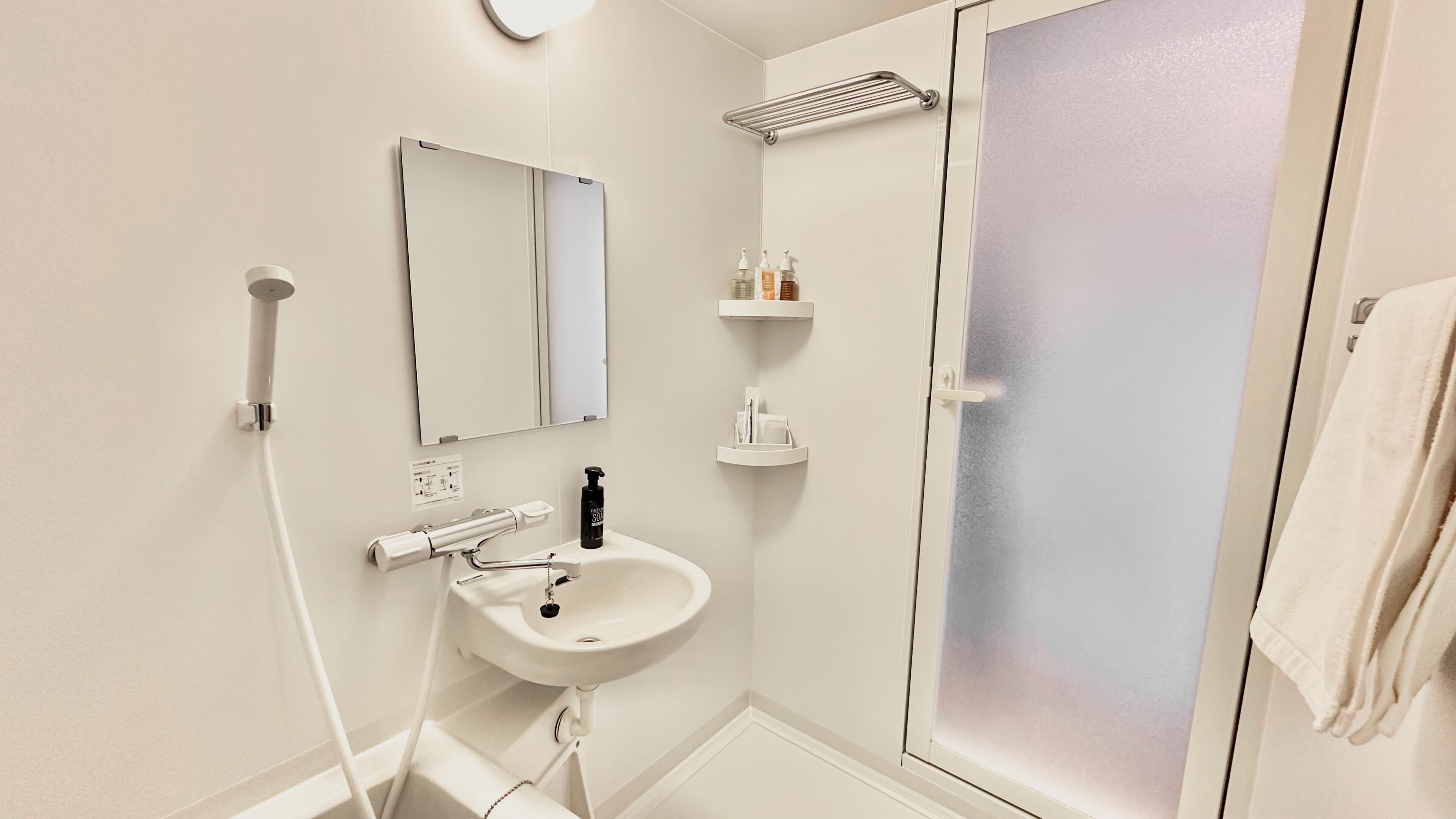 Une salle de bain avec un lavabo blanc, un miroir et une douche. On y trouve des articles de toilette sur une étagère et une serviette accrochée au mur.