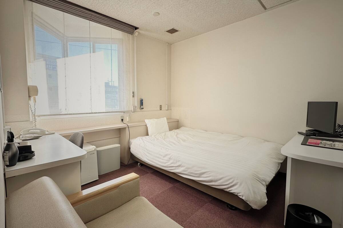 A hotel room with a bed, a desk, a chair, a TV, and a window with blinds.