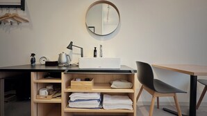 A modern hotel room with a wooden desk, a round mirror, a black chair, a white bedside table, and a shelf with towels and toiletries. - Hotel Sunroad (Amakusa)