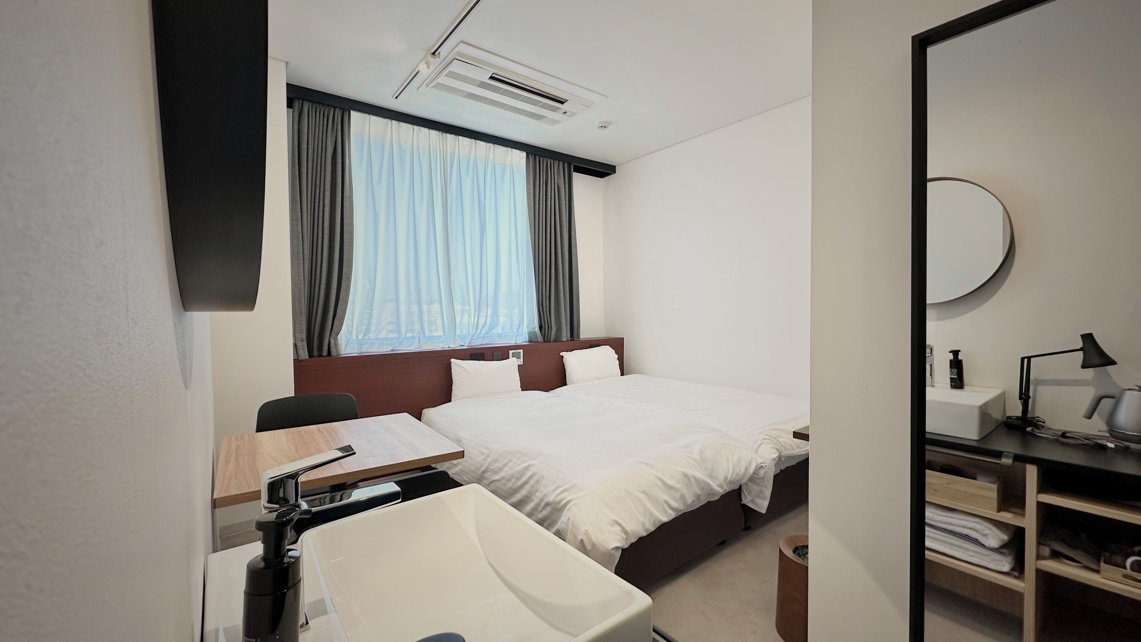 A hotel room with a bed, a desk with a chair, a sink, a mirror, and a window with curtains.