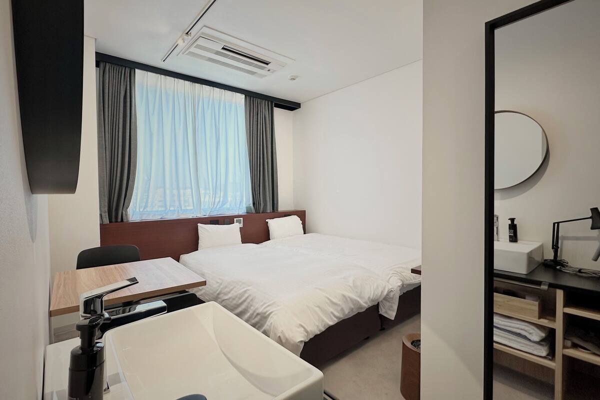 A hotel room with a bed, a desk with a chair, a sink, a mirror, and a window with curtains.