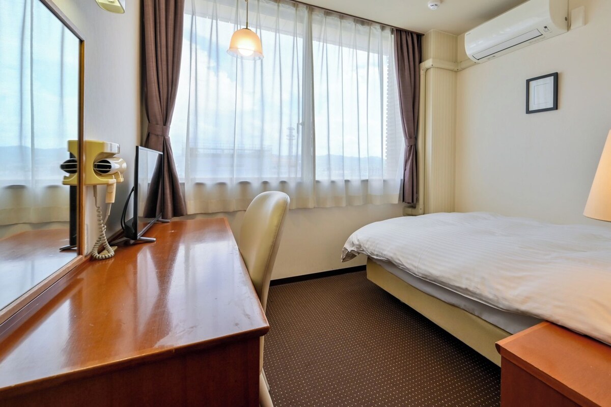 A hotel room with a bed, a desk with a telephone, a chair, a lamp, and a window with curtains.
