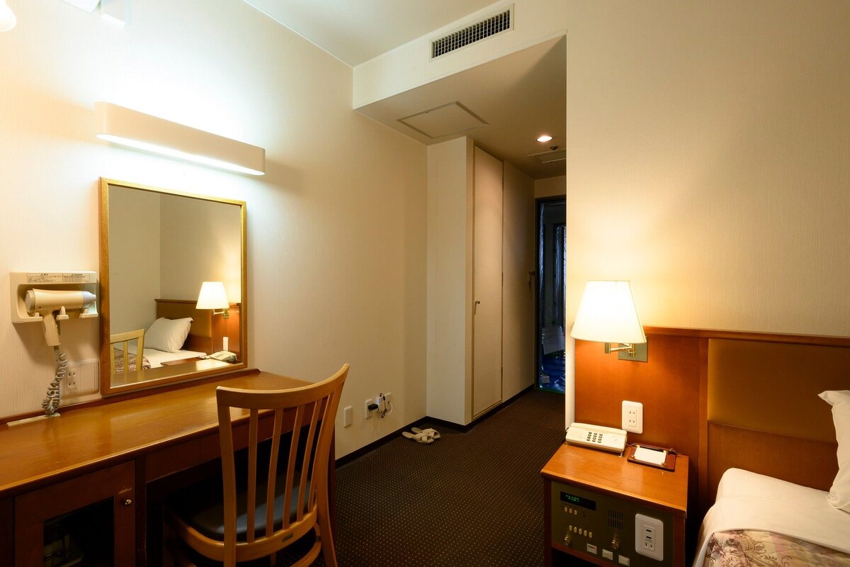 A hotel room with a desk, chair, bed, bedside table, lamp, and a mirror.