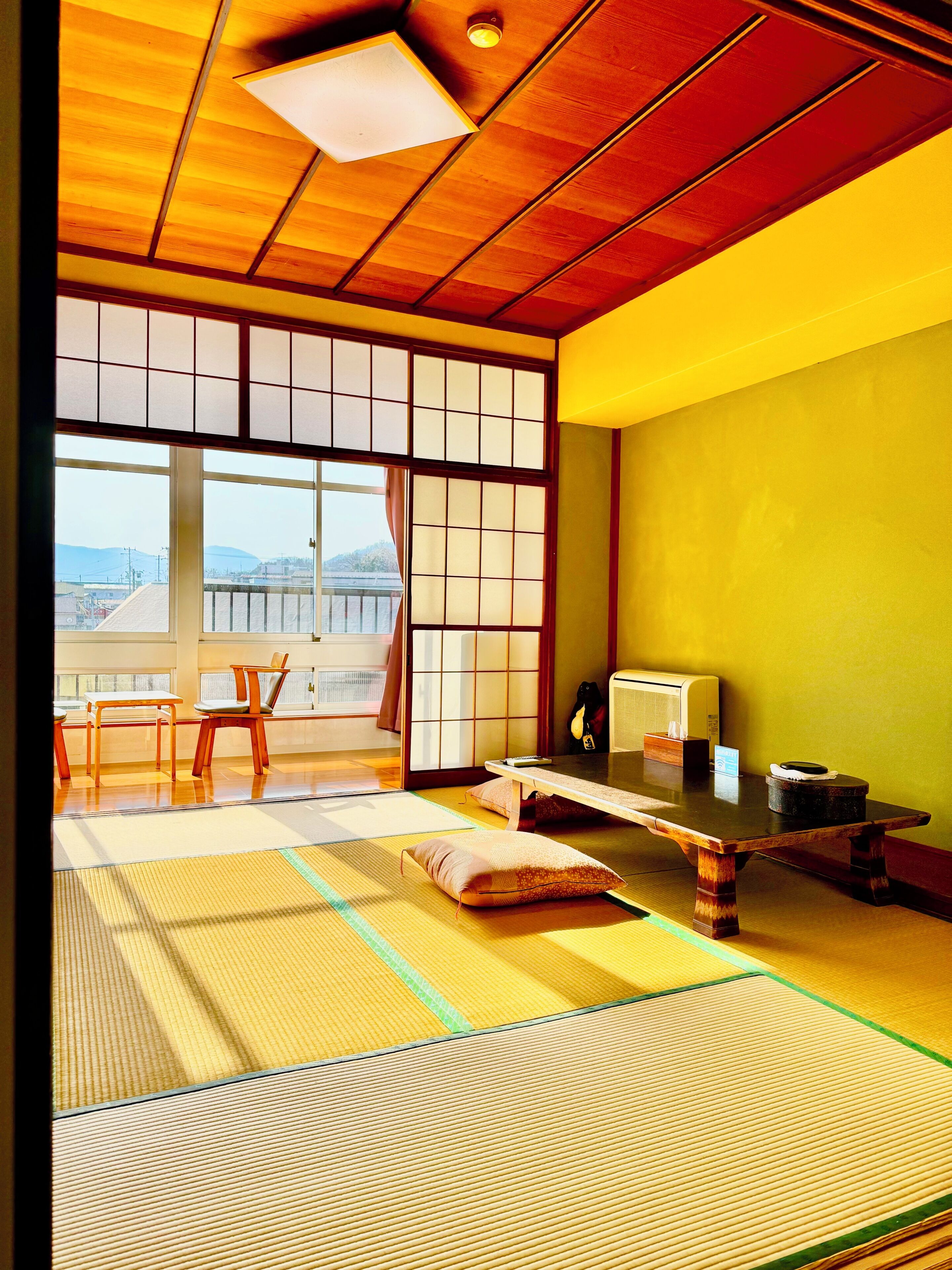 Ein traditioneller japanischer Raum mit Tatami-Böden, einem niedrigen Tisch, Stühlen, einer Schiebetür und Blick ins Freie.