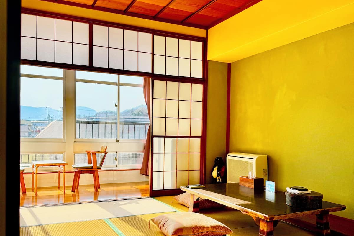 Una habitación japonesa tradicional con suelo de tatami, una mesa baja, sillas, una puerta corredera y vistas al exterior.