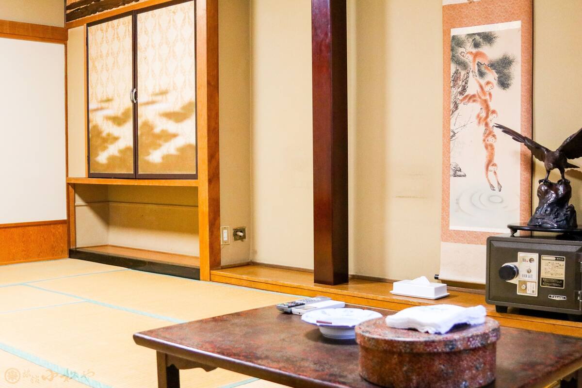 Una habitación japonesa tradicional con suelo de tatami, una mesa de madera con platos y una pequeña estatua, y una pintura en la pared.