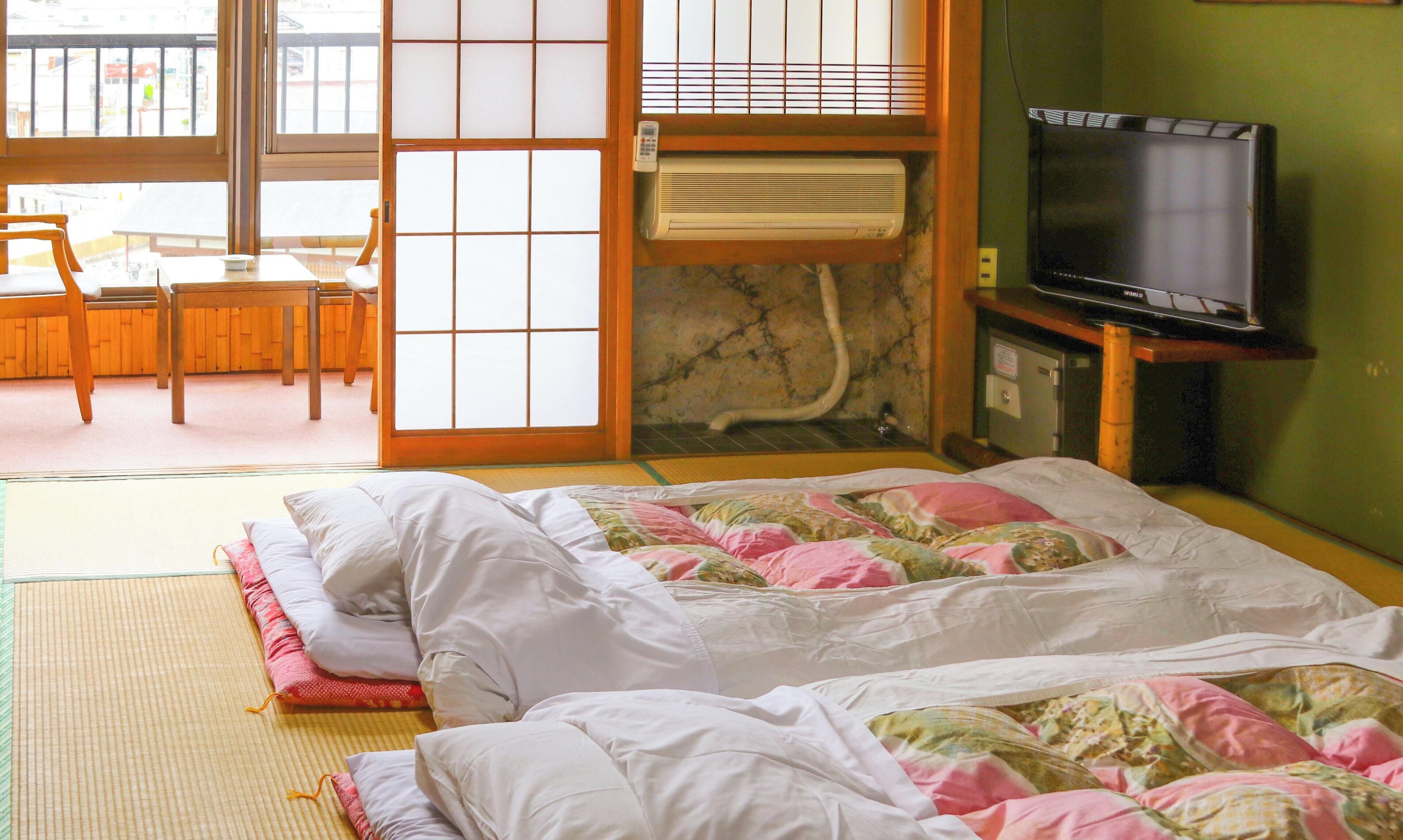Ein traditioneller japanischer Raum mit Tatami-Boden, einer Futon-Liege, einem Fernseher und einer Schiebetür zu einem Balkon.
