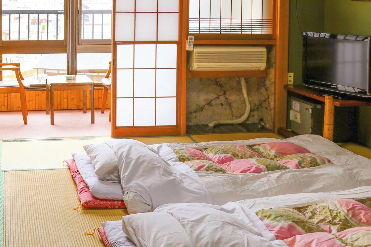 Una habitación japonesa tradicional con suelo de tatami, una cama futón, un televisor y una puerta corredera que da a un balcón.