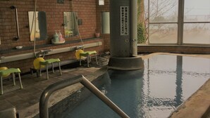 Une piscine intérieure avec un sol carrelé, quelques chaises vertes, une grande fenêtre et un mur en briques agrémenté d’un miroir et d’un porte-serviettes.