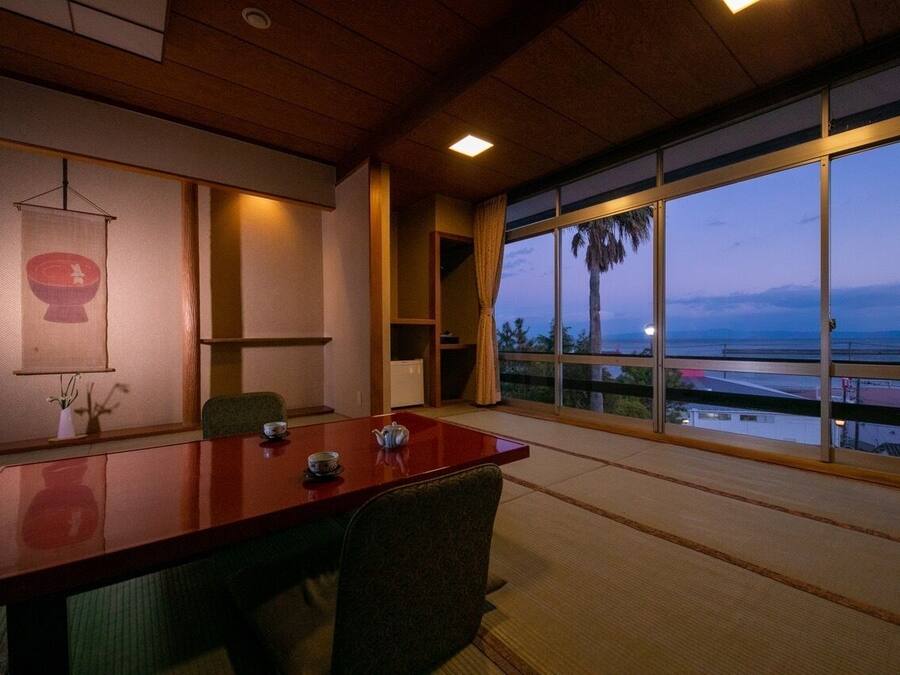 Een kamer met een houten plafond, een groot raam met uitzicht op palmbomen en een gebouw, een tatami vloer en een traditionele Japanse tafel met theeservies.