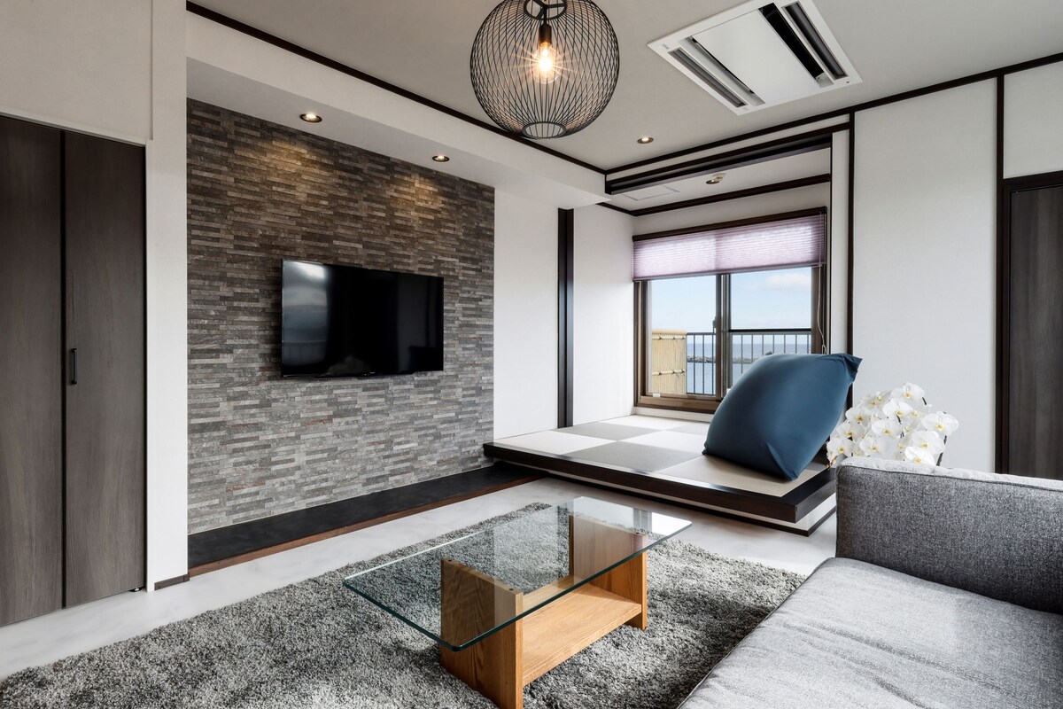 Una sala de estar moderna con un televisor de pantalla plana montado en una pared con textura, una mesa de centro de vidrio, un sofá y un ventanal con vistas.