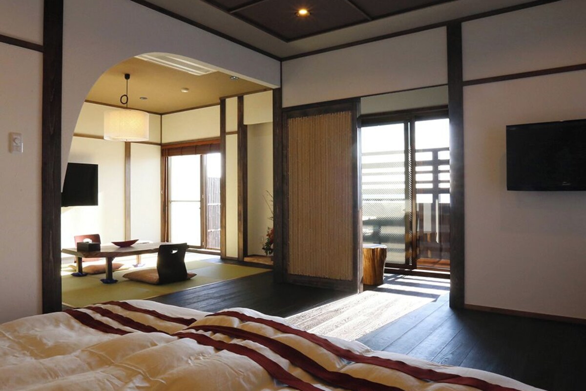 A spacious bedroom with a large bed, a television, and a view through a sliding door to a balcony.