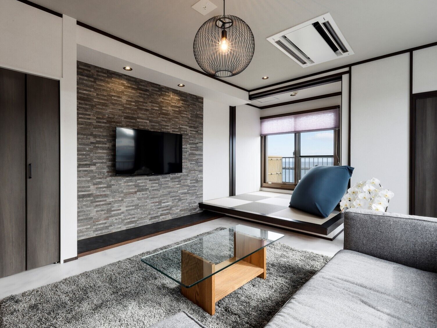 A modern living room with a flat-screen TV, a glass coffee table, a sofa, and a view of the outdoors.