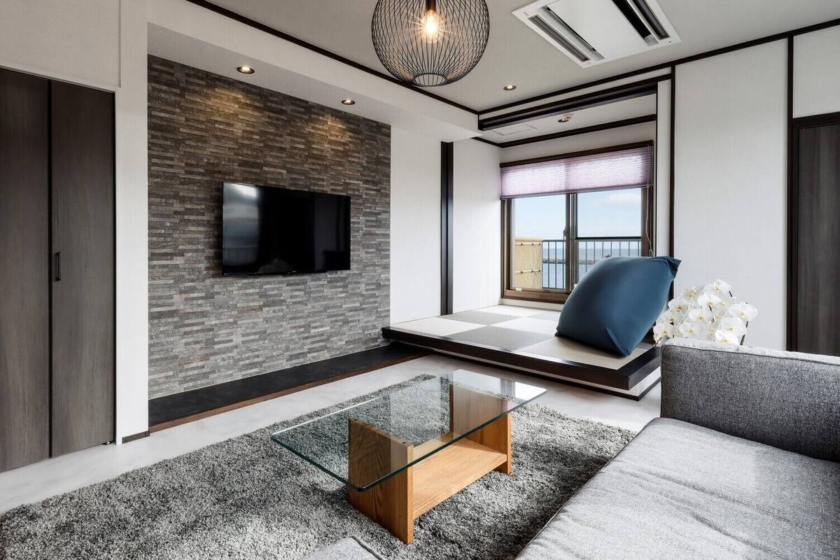 A modern living room with a flat-screen TV, a glass coffee table, a sofa, and a view of the outdoors.