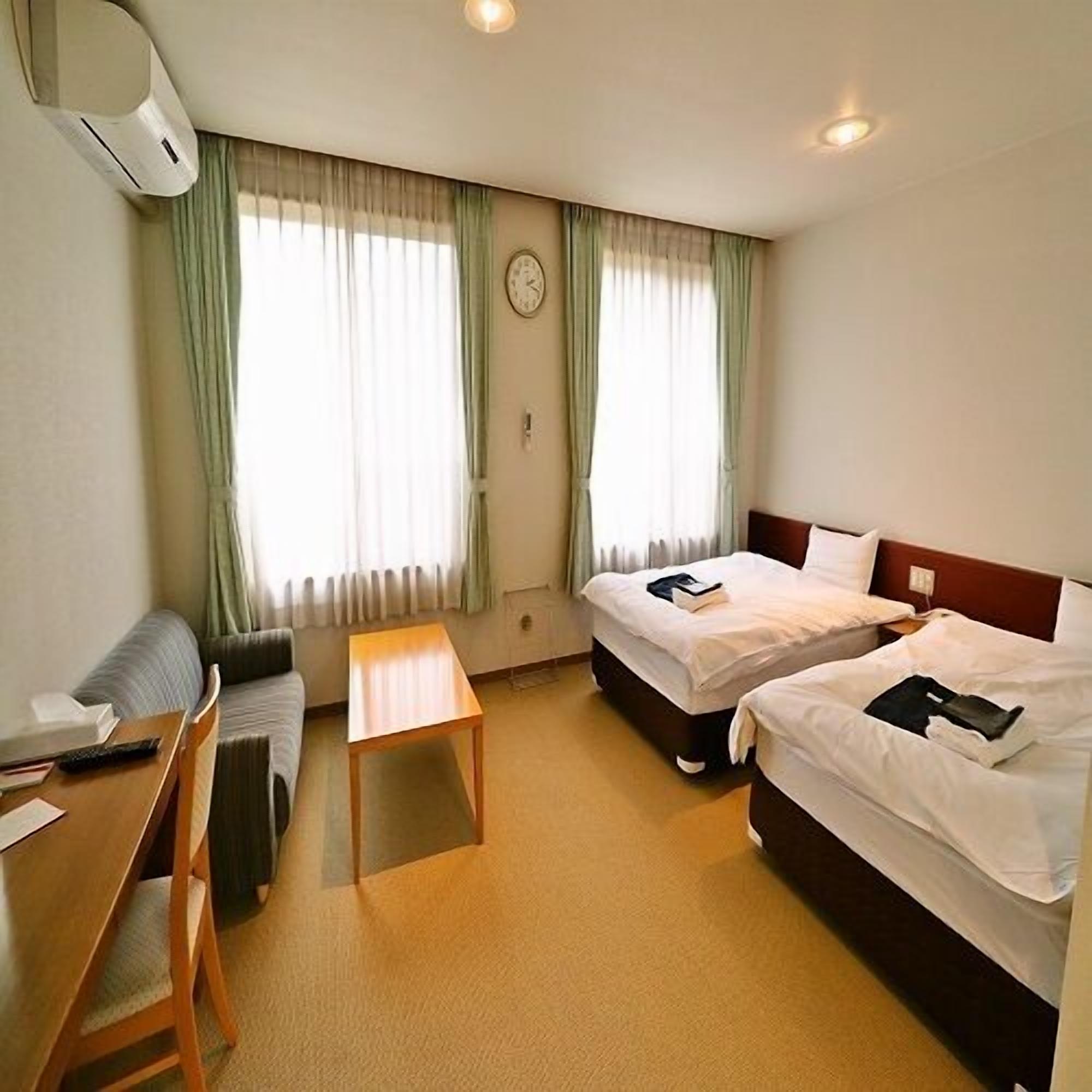 A hotel room with two beds, a desk, a chair, a small table, and a clock on the wall.