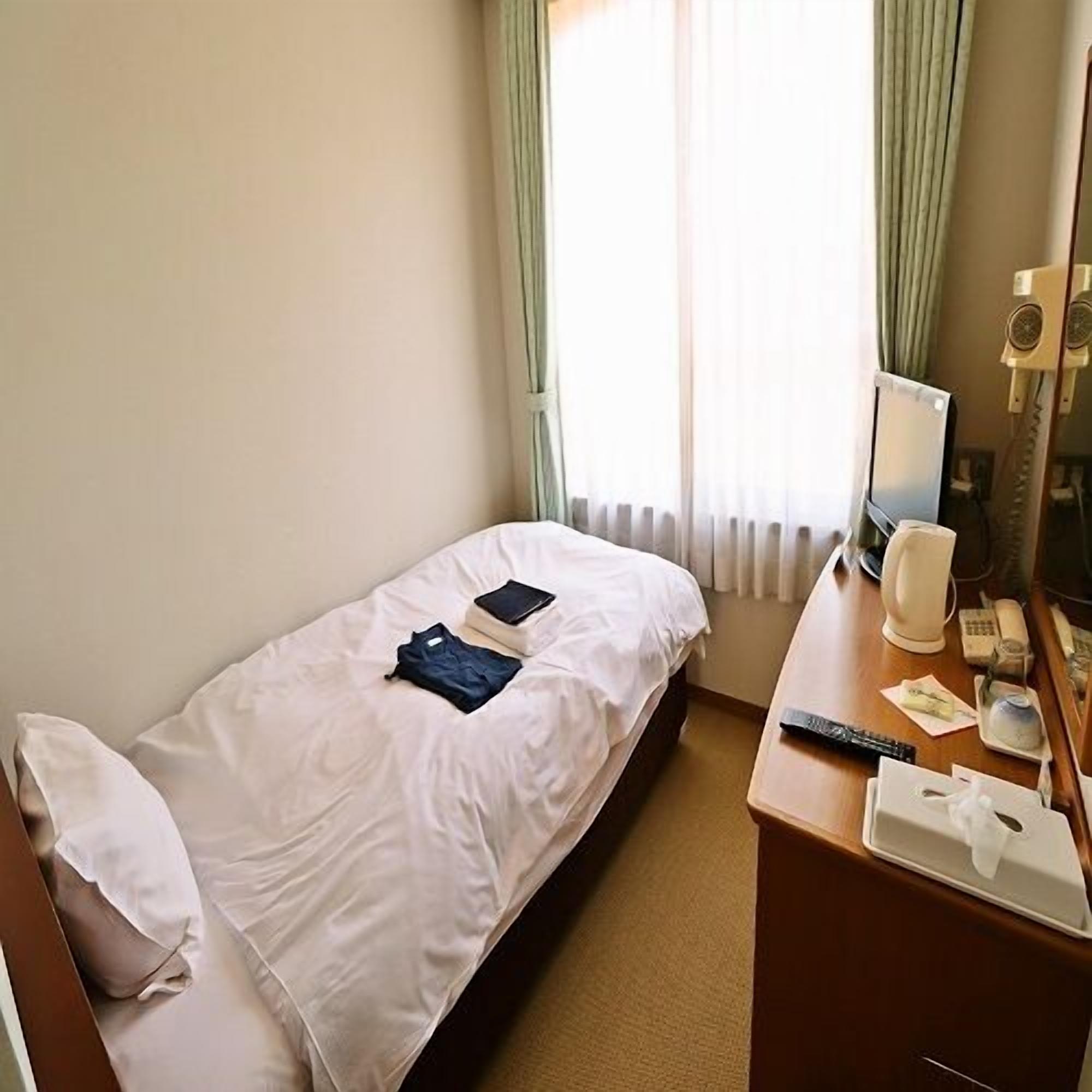 A single bed with a white sheet, a bedside table with a lamp, a mirror, and a TV.