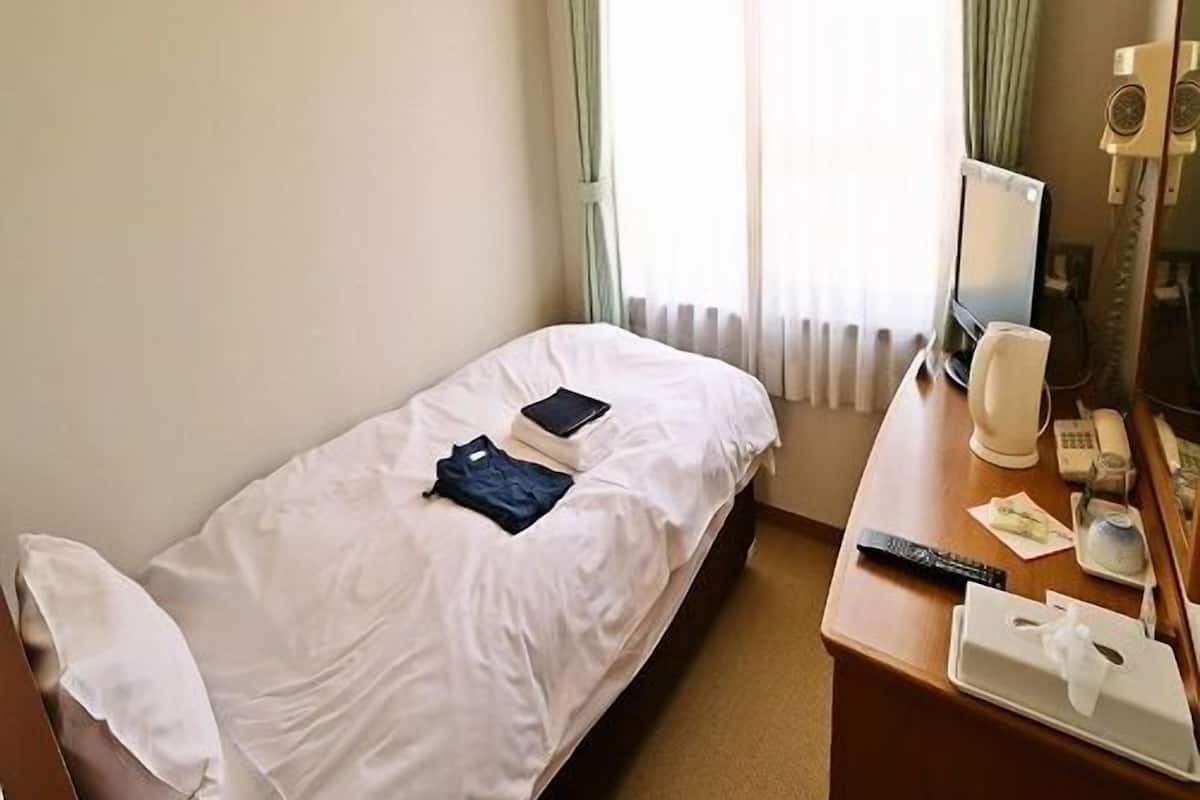 Un lit simple avec un drap blanc, une table de chevet avec une lampe, une télécommande et un miroir.