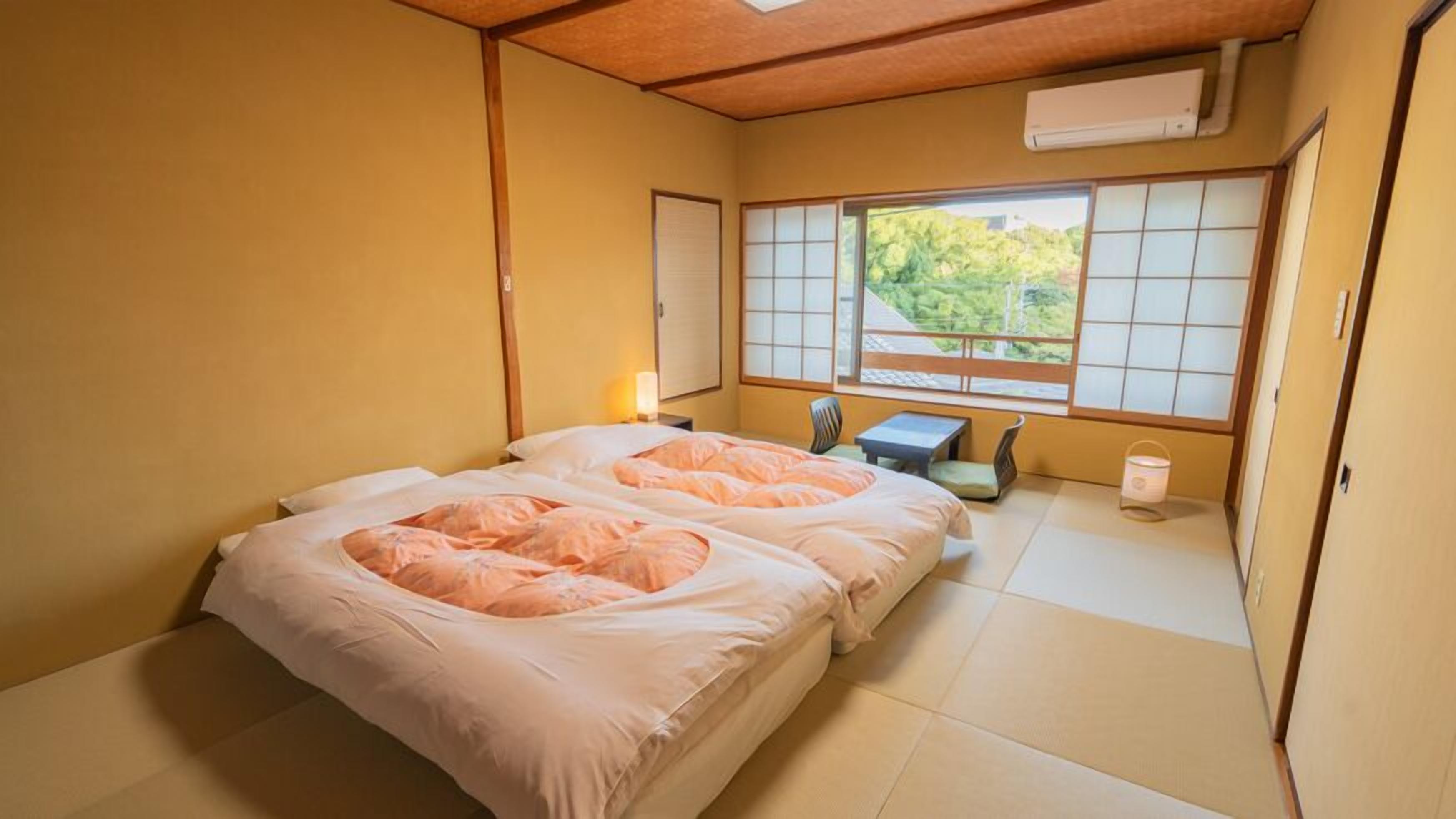A traditional Japanese-style bedroom with tatami flooring, a futon bed, a small table, and a window with a view of greenery.