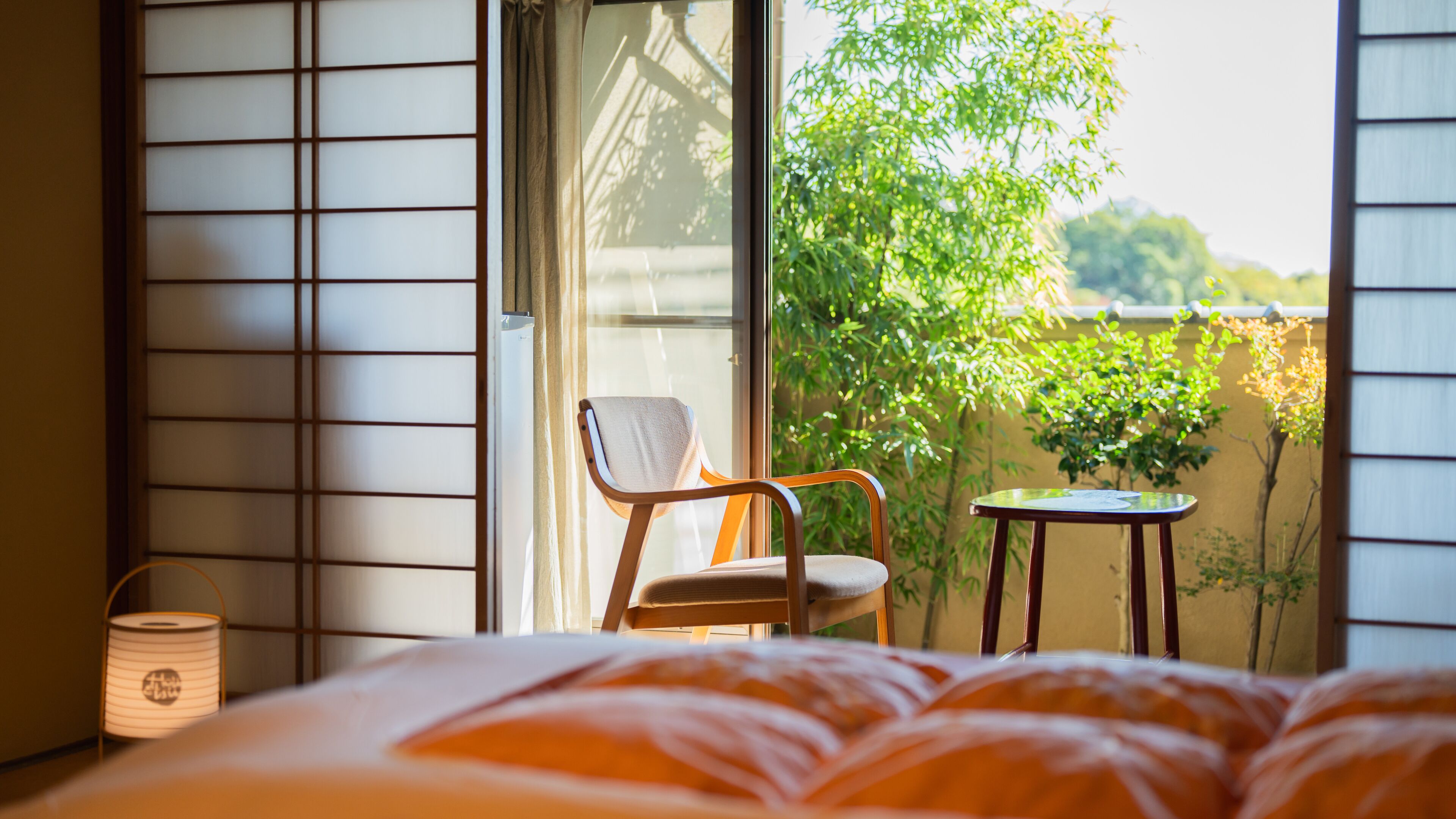 A room with a bed, a chair, a small table, a lamp, and a view of greenery outside.