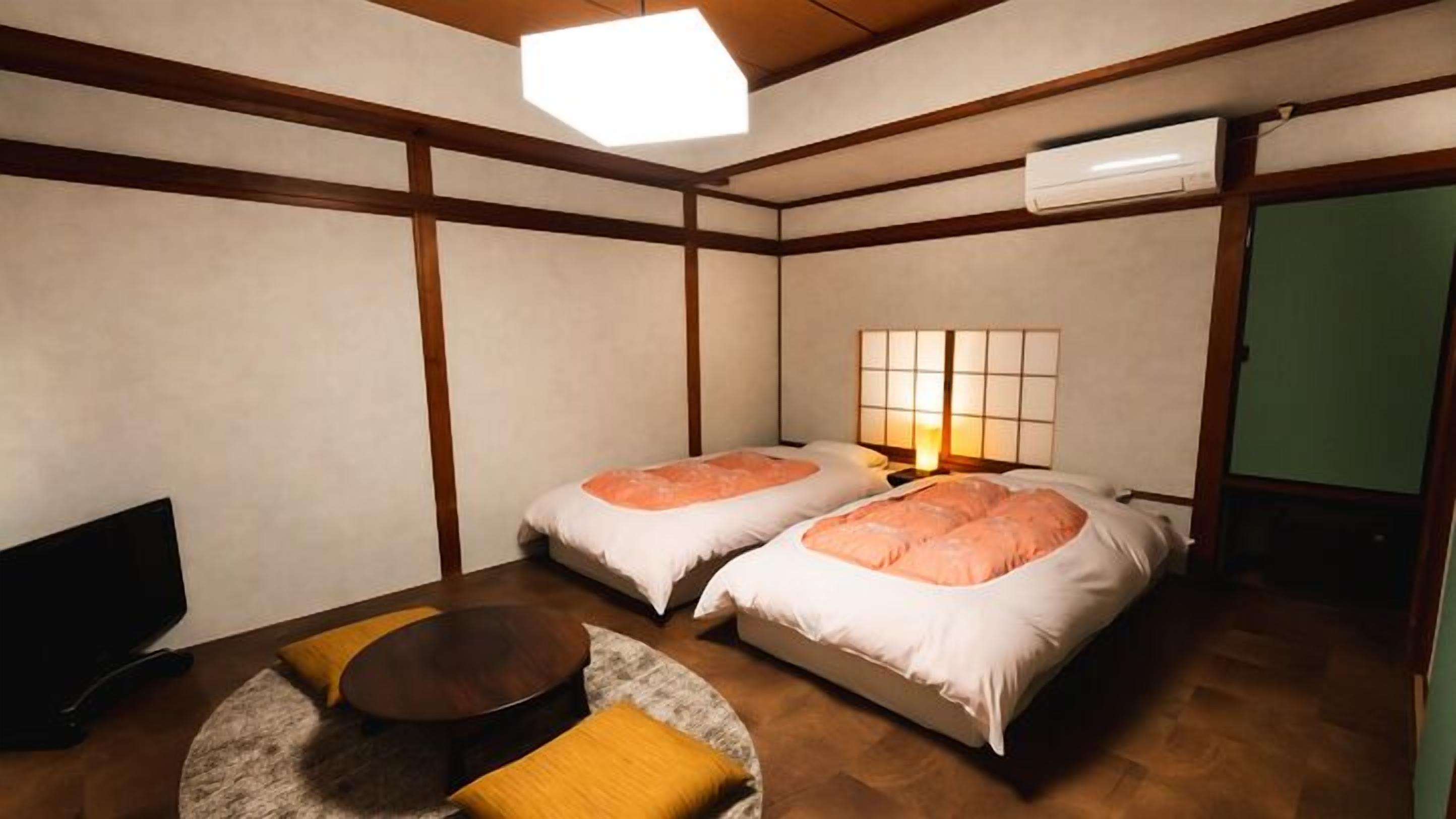 A room with two beds, a small round table, a sofa, a ceiling light, and an air conditioner.