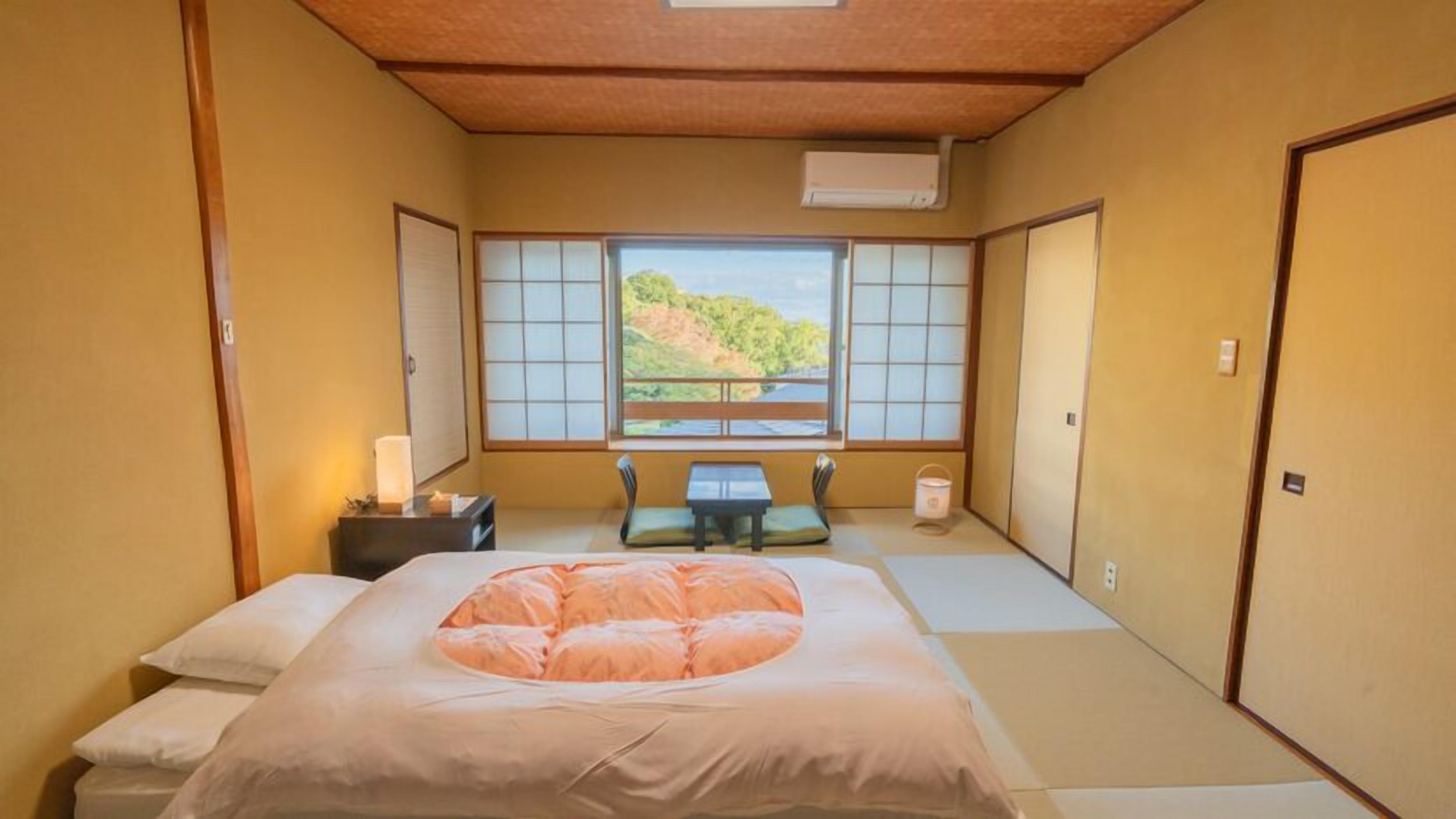 A traditional Japanese-style room with tatami flooring, a futon bed, a small table with a cushion, and a window with a view of a hillside.