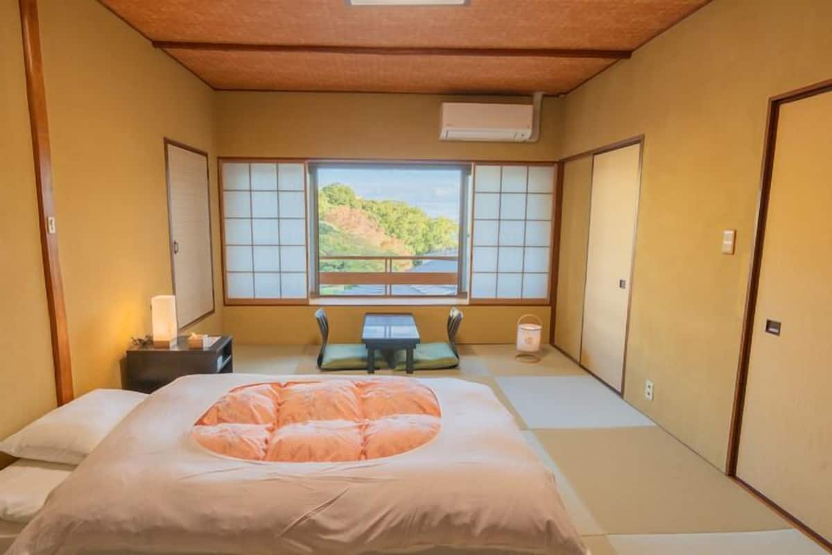 A traditional Japanese-style room with tatami flooring, a futon bed, a small table with a cushion, and a window with a view of a hillside.