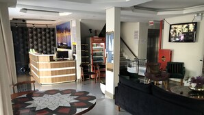 En moderne hotellobby med en receptionsdisk, et siddeområde med et rundt bord, en sofa og en trappe til en etage ovenover.
