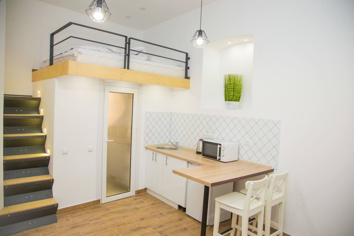 Eine kompakte Küche mit Mikrowelle, Spüle und einem Holztisch. Eine Treppe mit Metallgeländer führt zu einem Schlafbereich als Erker.