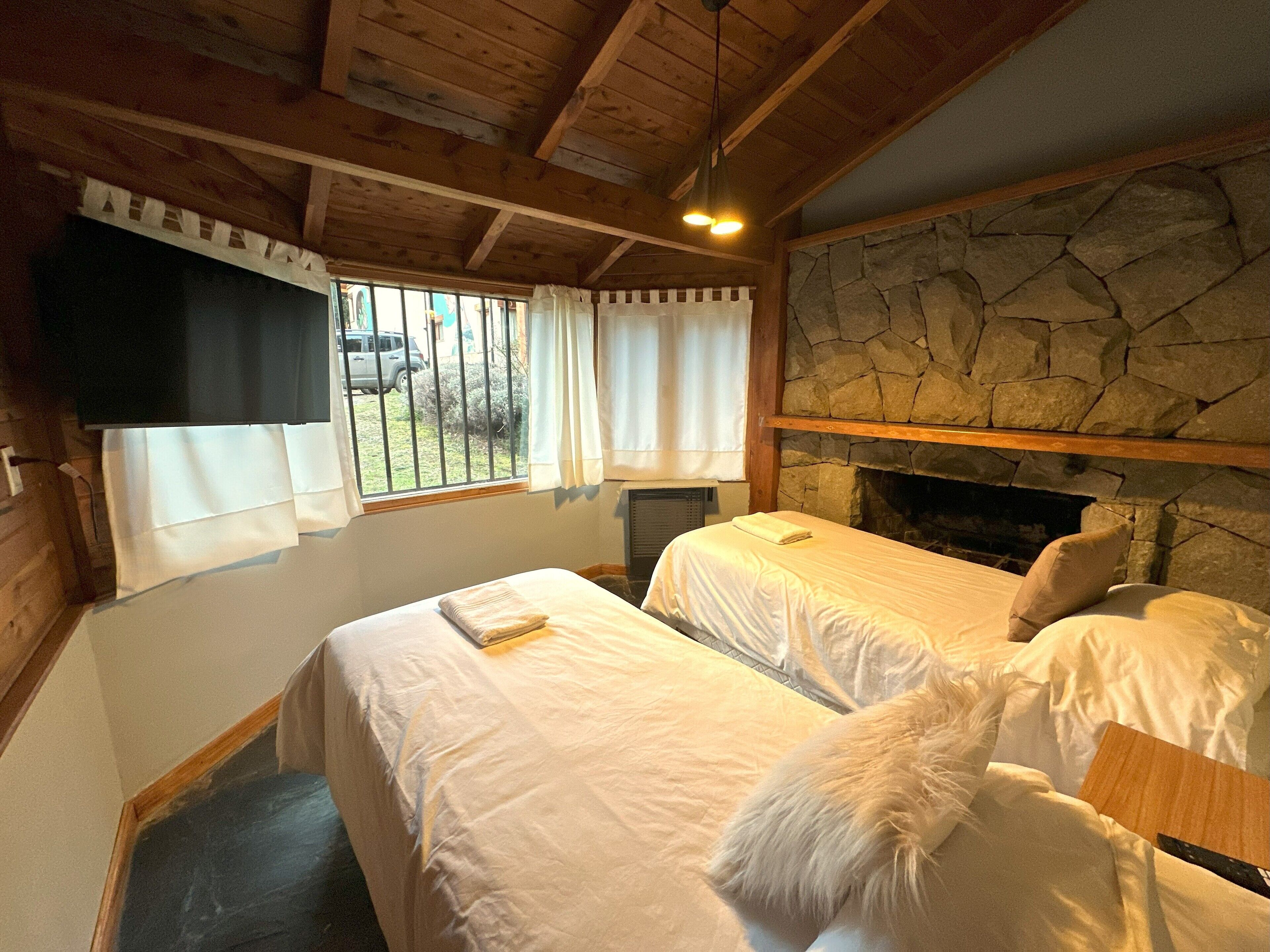 A room with two beds, a stone fireplace, wooden beams, a window with curtains, and a view of the outside.