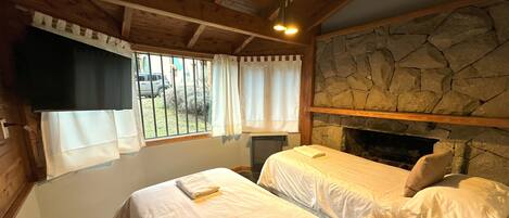 A room with two beds, a stone fireplace, wooden beams, a window with curtains, and a view of the outside.