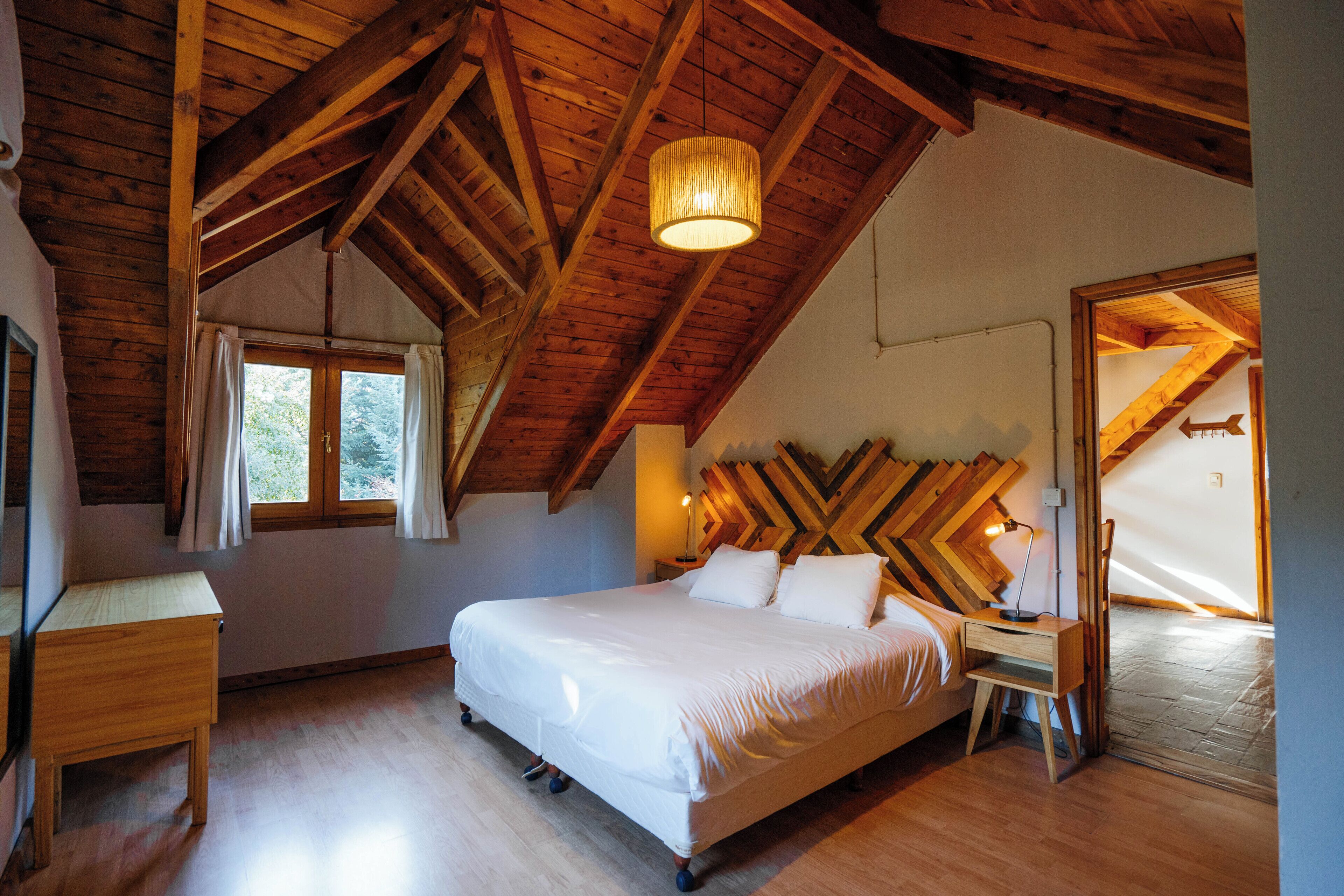 A bedroom with a wooden ceiling, a bed with white bedding, a bedside table, and a window with curtains.
