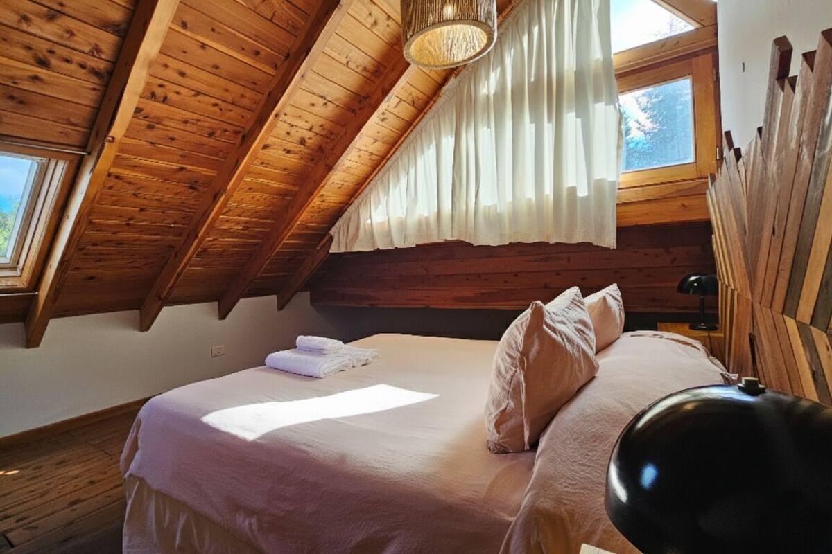 Une chambre avec un plafond en pente, des murs en bois, un lit avec des oreillers et une couverture, une table de chevet et une fenêtre avec des rideaux.